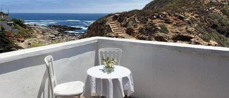 A balcony with a white table and chair, overlooking a rocky coastline and the ocean.