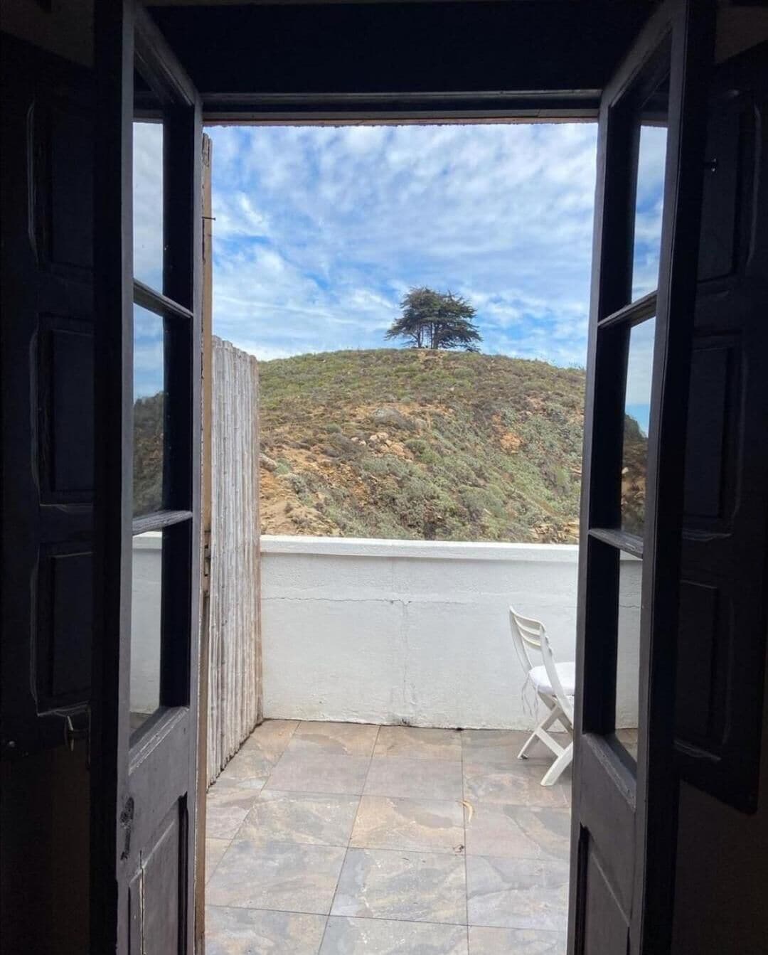 An open door leading to a balcony with a view of a hill and a tree.