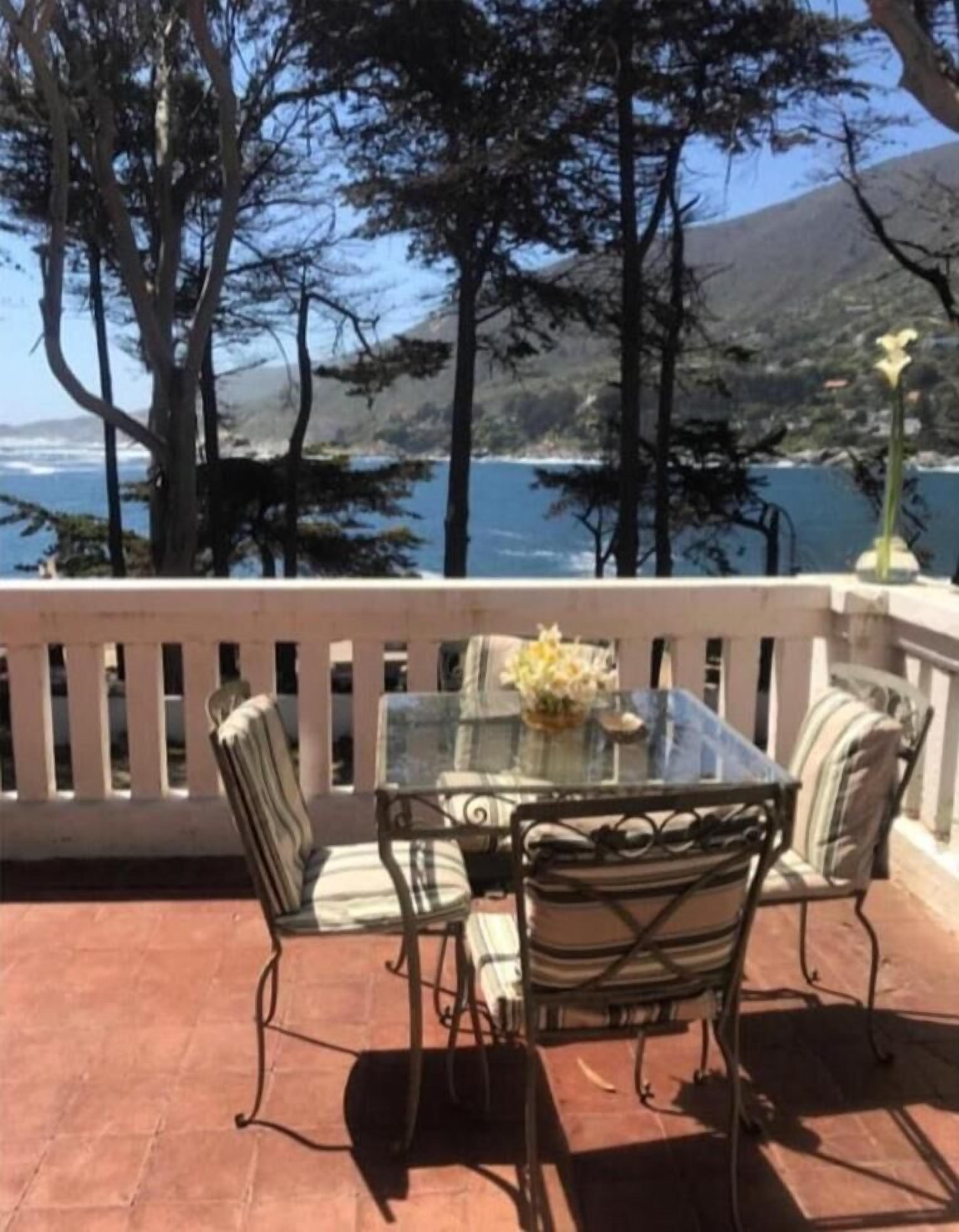 Una terraza con mesa y sillas de vidrio, con vista a un paisaje costero con árboles y al mar.