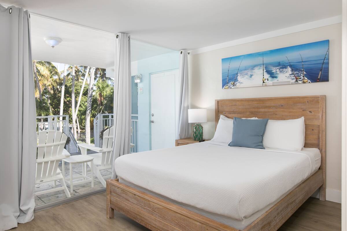 A bedroom with a wooden bed, white bedding, a painting of fishing boats, and a view of palm trees through the door.