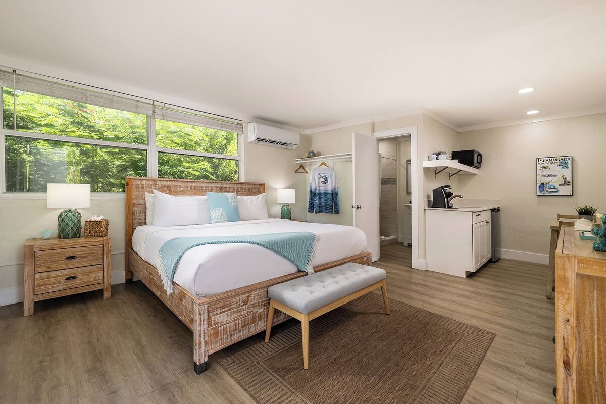 A bedroom with a large bed, bedside tables, a bench, a small kitchenette, and a bathroom.