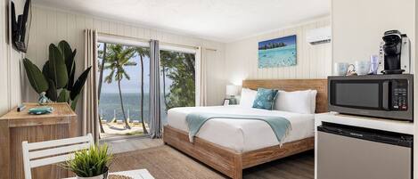 A bedroom with a bed, a desk, a dining table, a plant, a view of the ocean, and a kitchen area with a microwave.