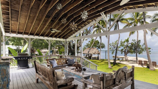 A wooden deck with rustic furniture, a grill, and a view of the beach and palm trees. - La Jolla Resort (Islamorada)