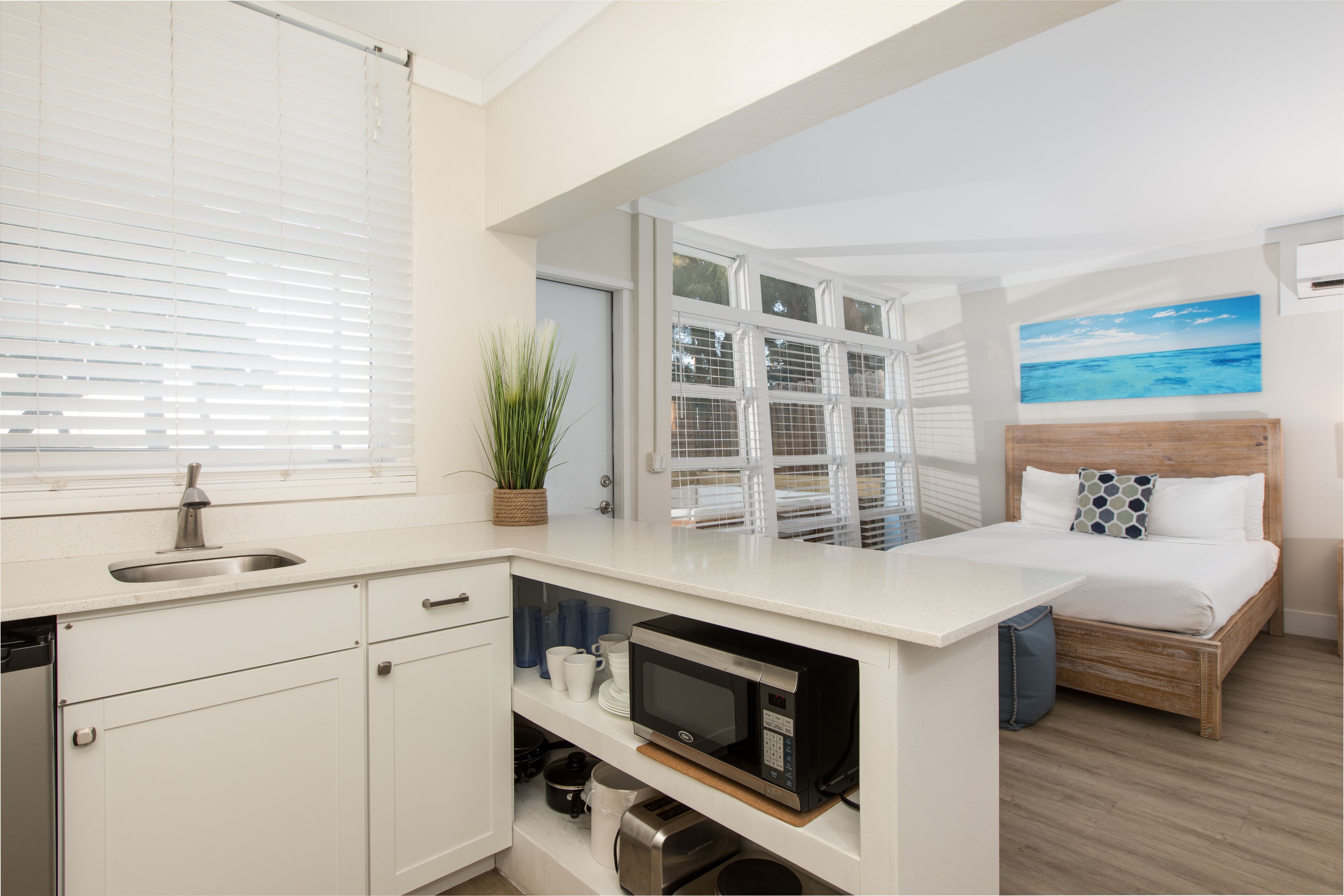 A compact hotel room with a kitchenette, a bed with a wooden headboard, a painting of the ocean, and a potted plant.