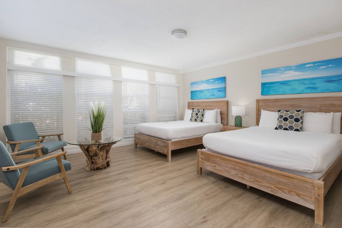A hotel room with two beds, a wooden coffee table, a chair, and large windows with blinds.
