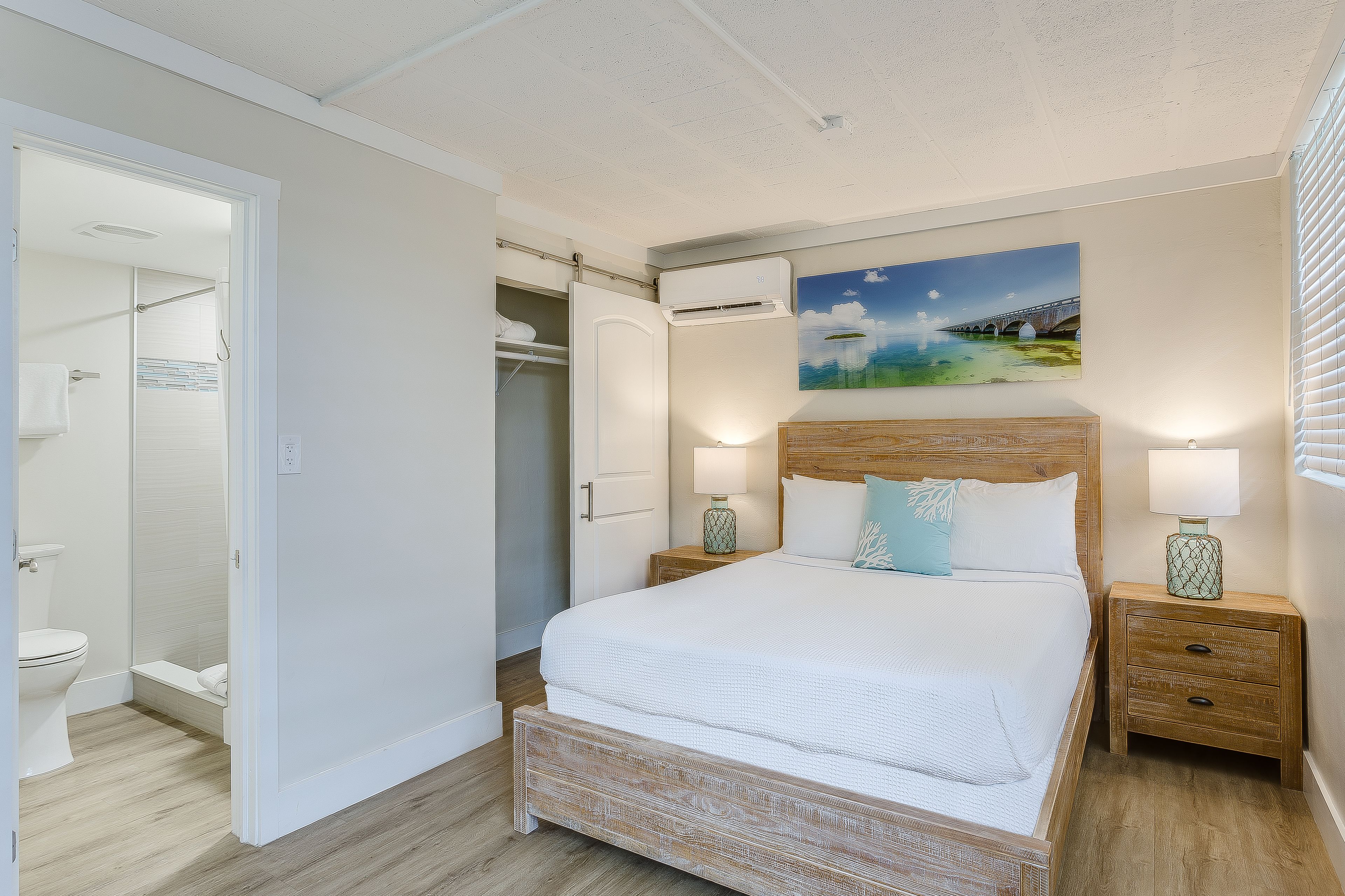 Un dormitorio con una cama grande, un cabecero de madera, dos mesitas de noche con lámparas, un cuadro en la pared y un armario abierto.