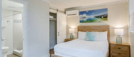 Un dormitorio con una cama grande, un cabecero de madera, dos mesitas de noche con lámparas, un cuadro en la pared y un armario abierto.
