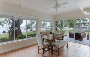 Un comedor con una mesa y sillas, ventanas amplias con vista a la playa y al océano, y un ventilador de techo.