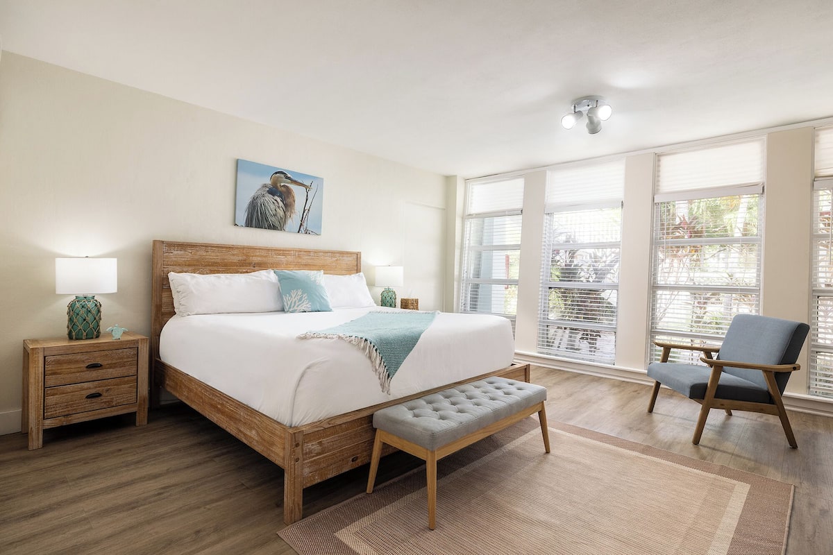 A bedroom with a wooden bed, a bench, a chair, and a large window with blinds.