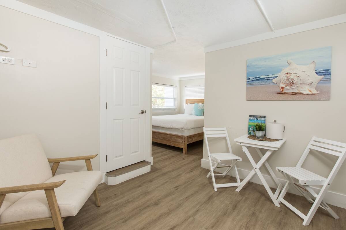 A bedroom with a wooden floor, a white bed, a white chair, a small table with a plant, and a painting of a seashell on the wall.