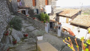 Un estrecho sendero de piedra que conduce a un edificio rústico de piedra con tejas de terracota, plantas en macetas y un pequeño jardín.
