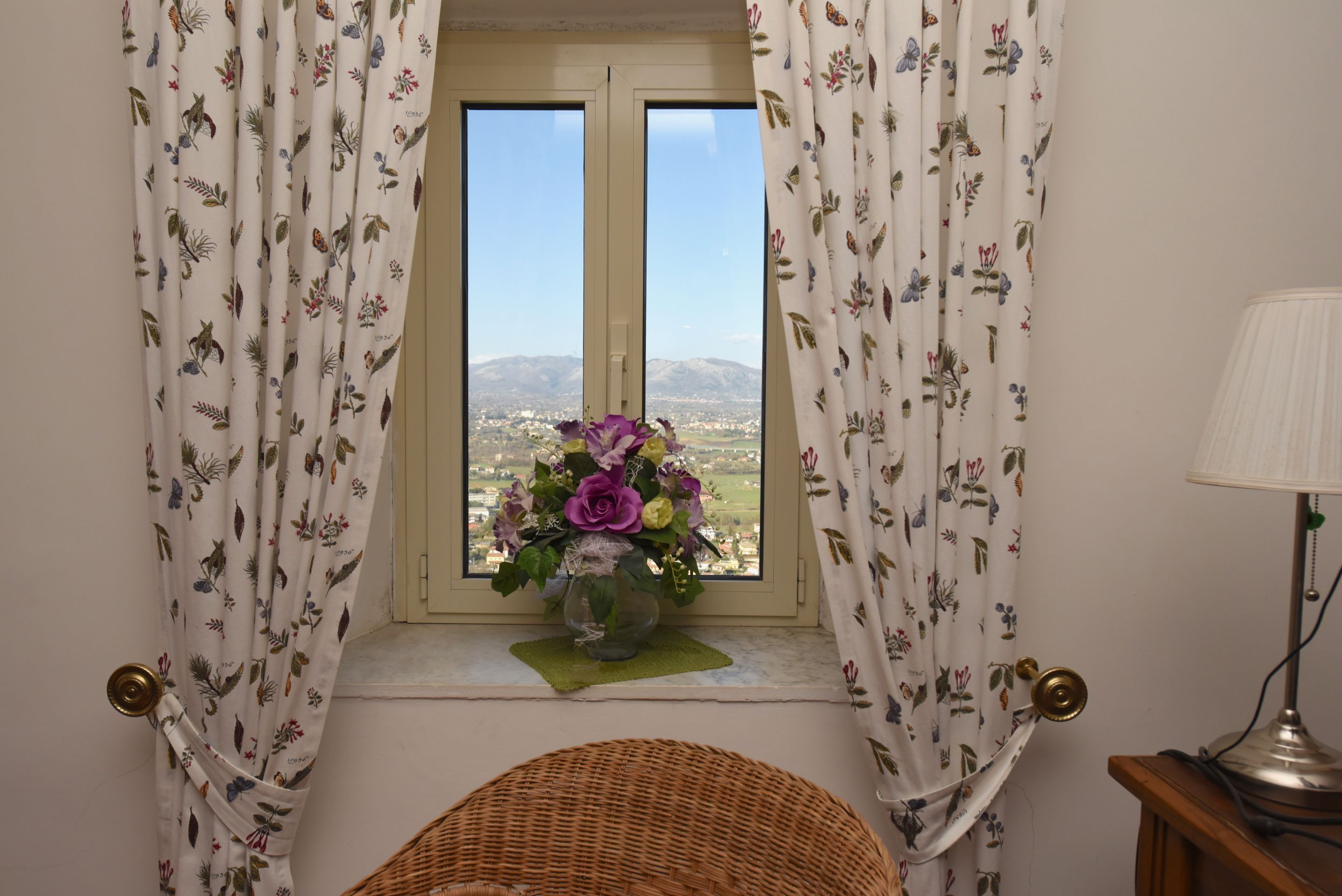 Una habitación con ventana, cortinas con motivos florales, una silla de mimbre y un florero con flores en el alféizar.