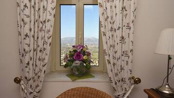 Una habitación con ventana, cortinas con motivos florales, una silla de mimbre y un florero con flores en el alféizar.