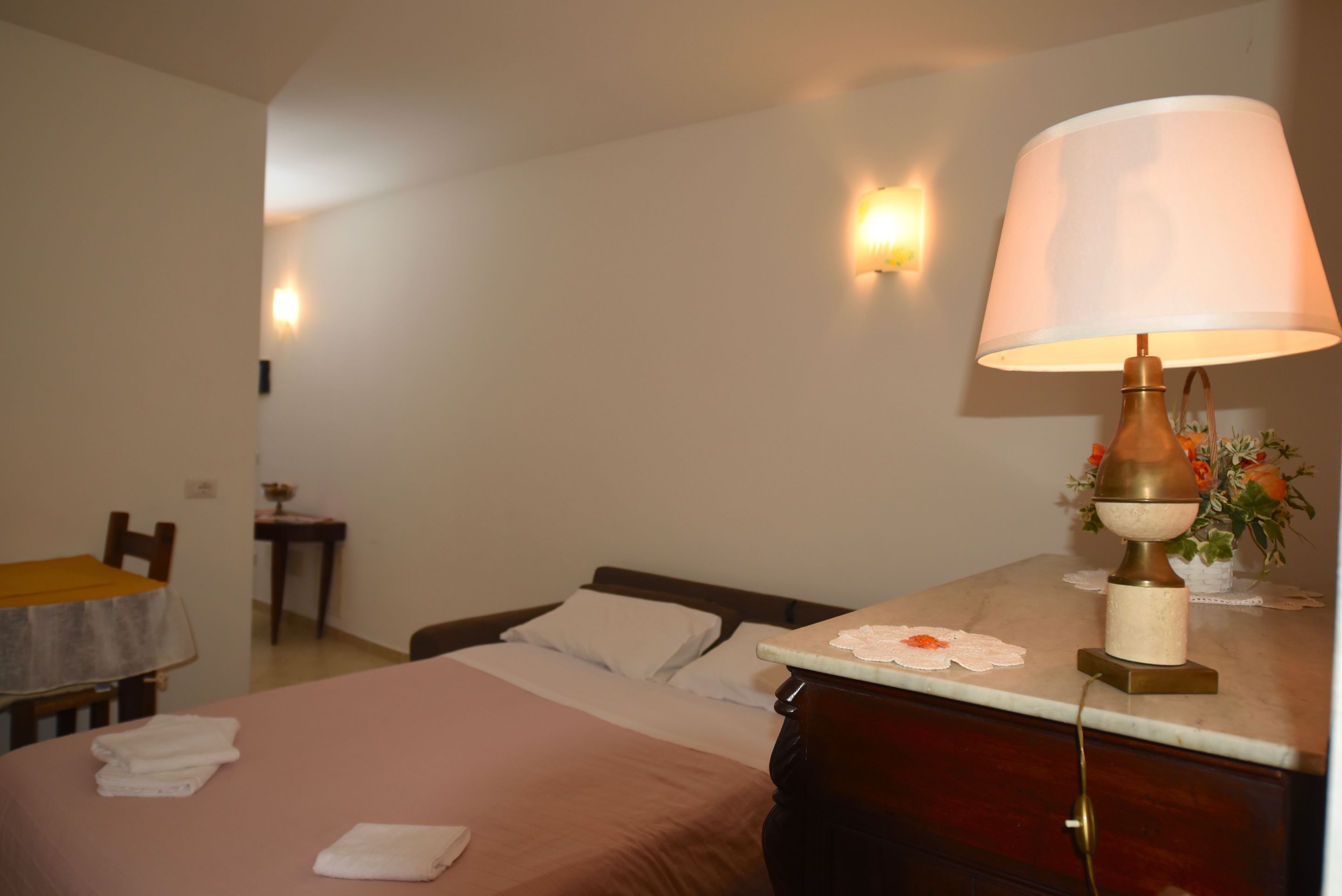 Habitación de hotel con cama, mesita de noche, lámpara y una pequeña mesa con un florero.