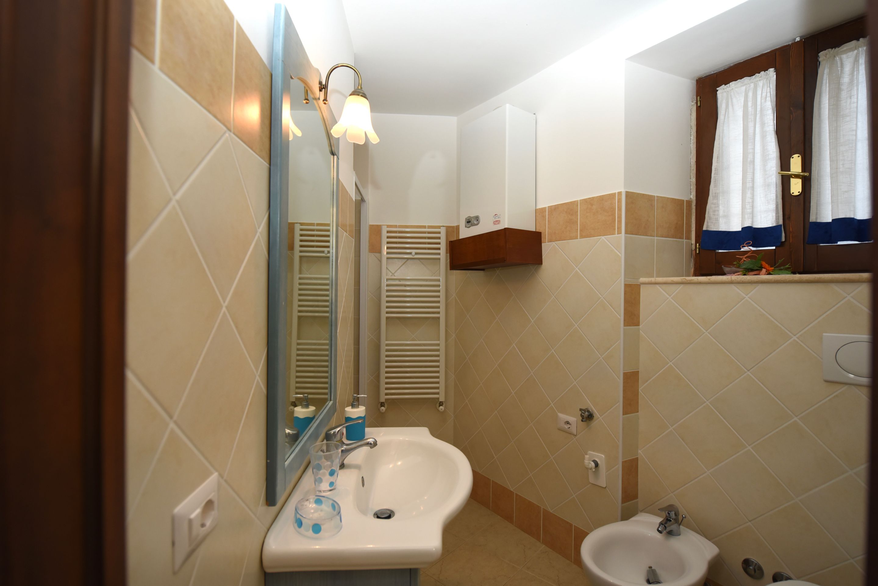 Une salle de bain équipée d’un lavabo, d’un bidet, d’un miroir, d’un porte-serviettes et d’une fenêtre avec des rideaux.