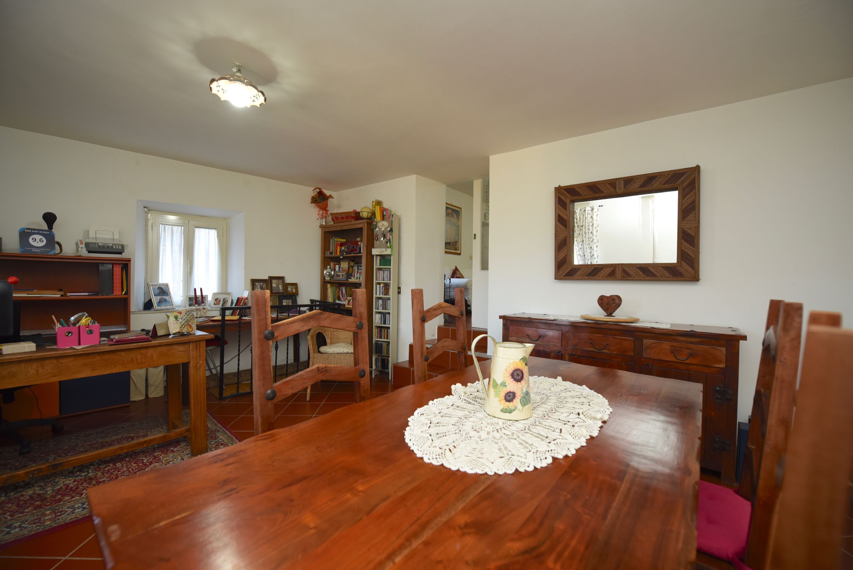 A dining room with a wooden table, chairs, a pitcher, and a bookshelf.