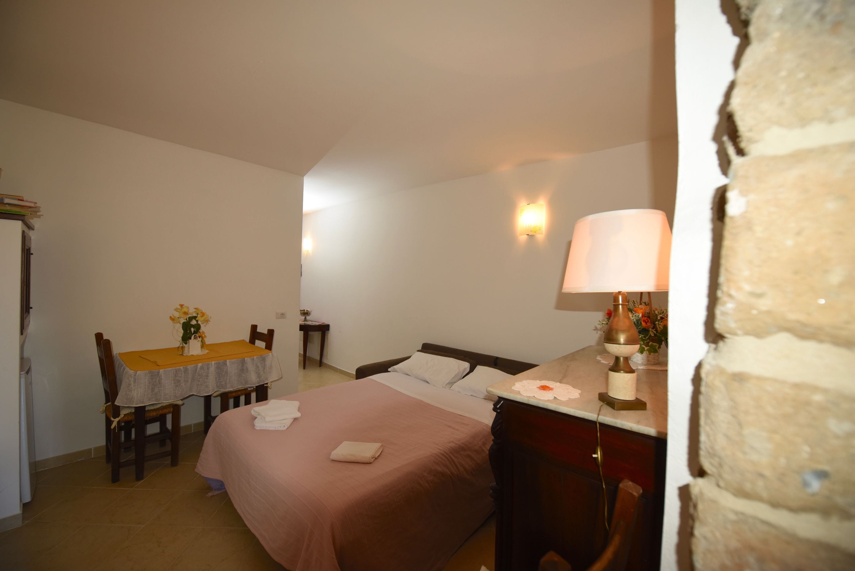 A bedroom with a bed, a bedside table with a lamp, a chair, a small table with a vase of flowers, and a wall-mounted light fixture.