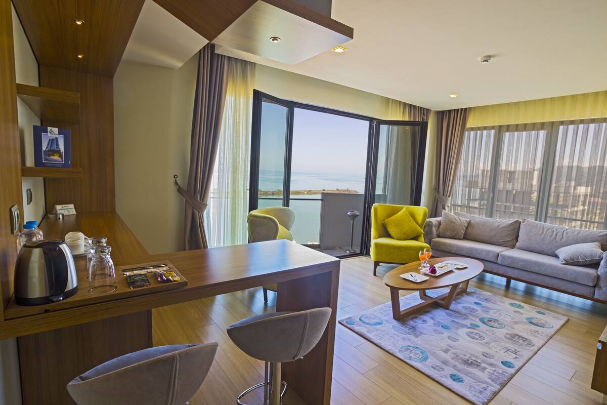 A modern hotel room with a wooden desk, a yellow armchair, a sofa, and a large window offering a view of the sea.