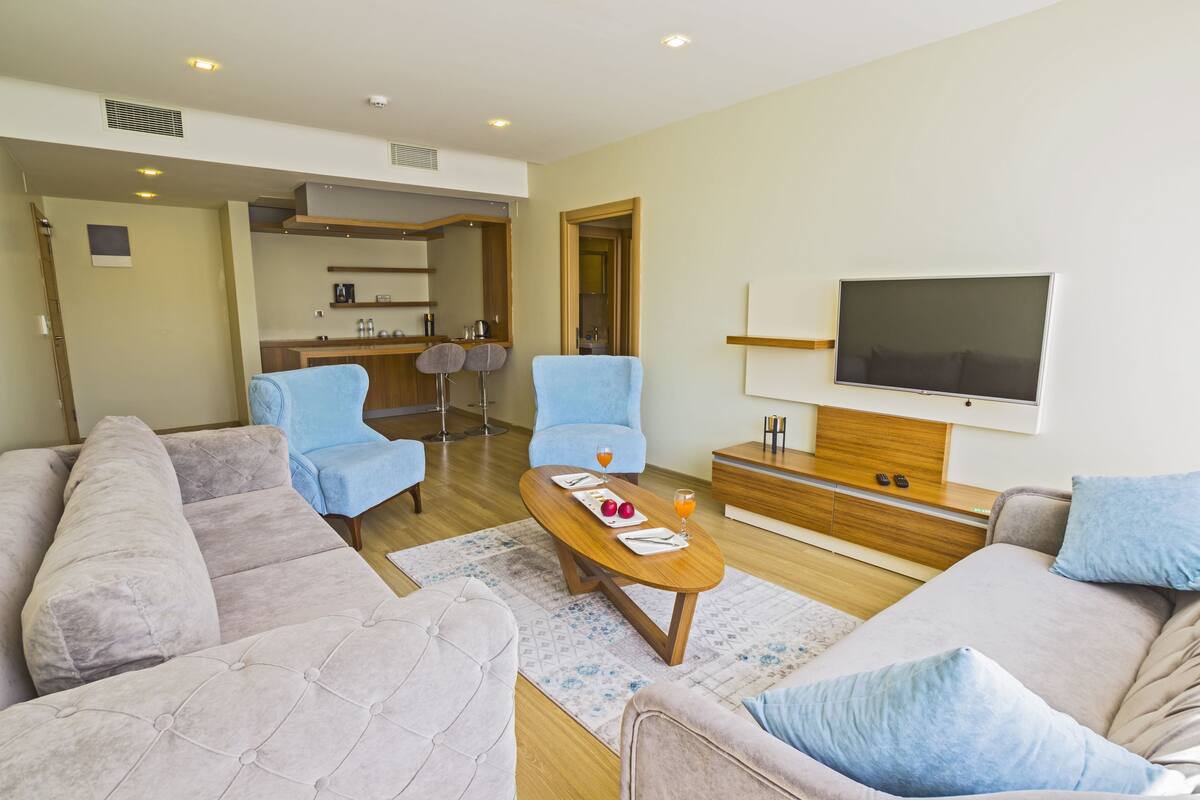 A living room with a sofa, two armchairs, a coffee table with fruit and a drink, a TV, and a kitchen in the background.