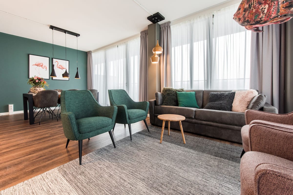 Een moderne woonkamer met een groene bank, twee groene fauteuils, een ronde houten salontafel en een grijze bank met een zachte plaid.