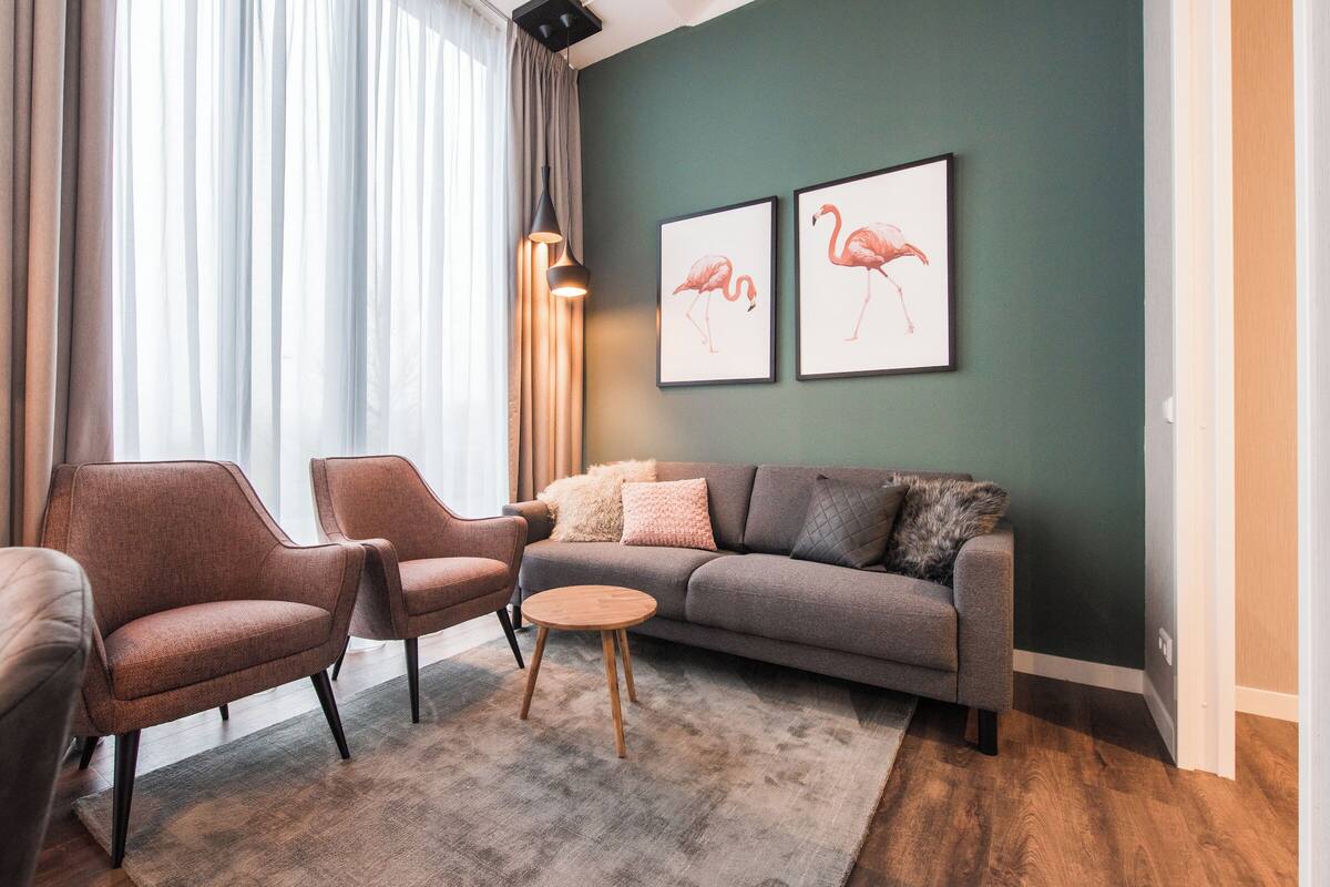 Een moderne woonkamer met een grijze bank, twee fauteuils, een ronde houten salontafel en een groot raam met lichte gordijnen.