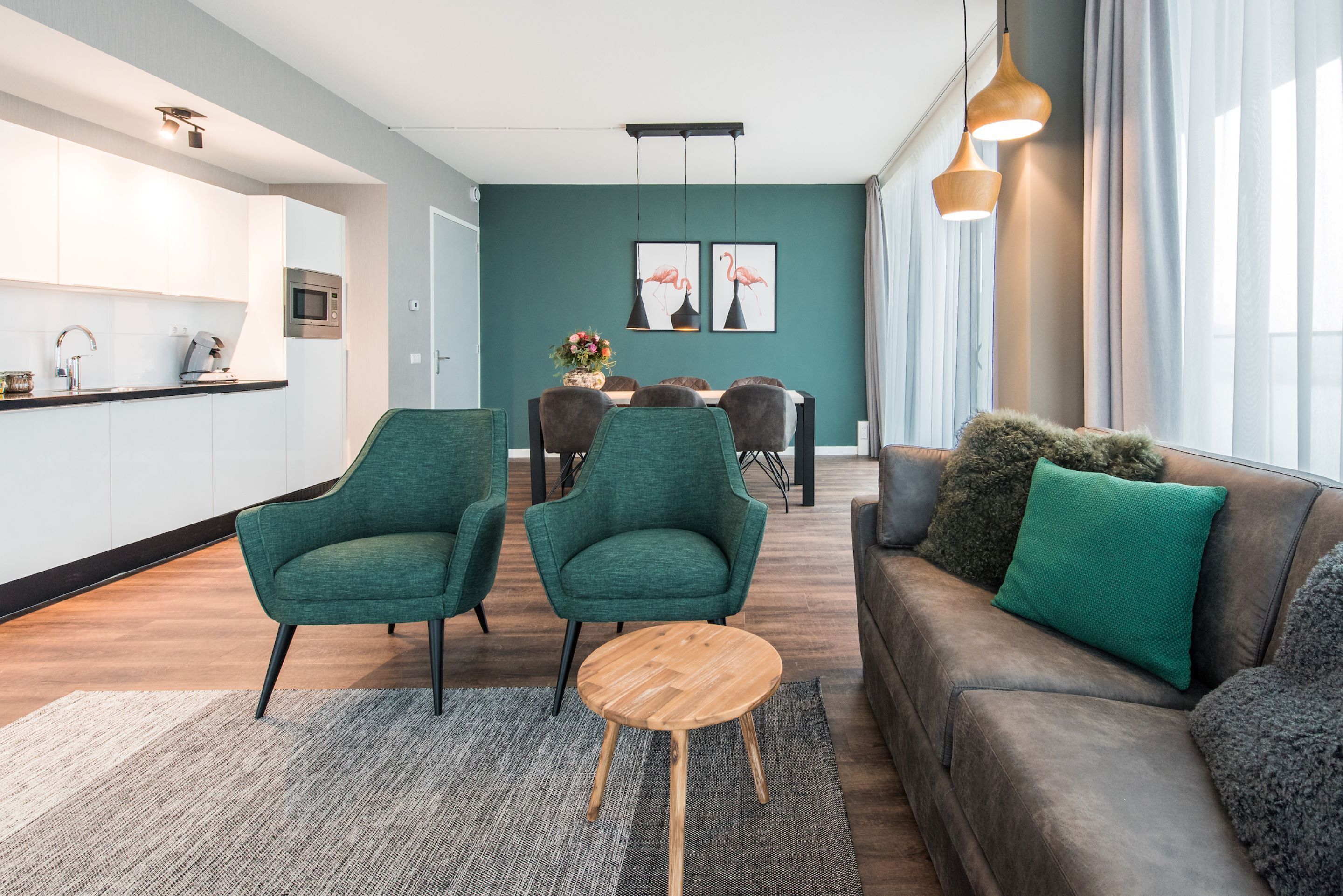 A modern living room with a brown leather sofa, two green armchairs, and a wooden coffee table. A dining area with a table and chairs is visible in the background.