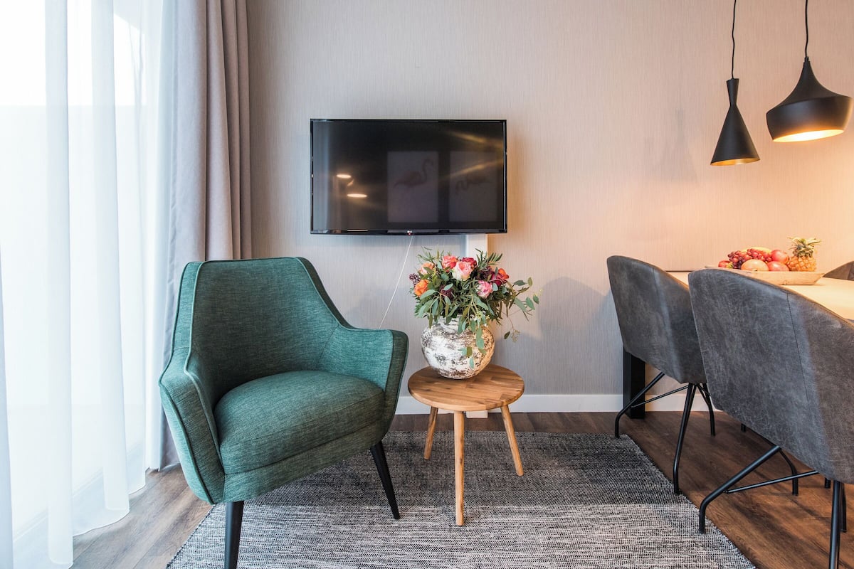Een moderne woonkamer met een groene fauteuil, een klein houten tafeltje met een bloevas, een eettafel met stoelen, en een flatscreen televisie aan de muur.