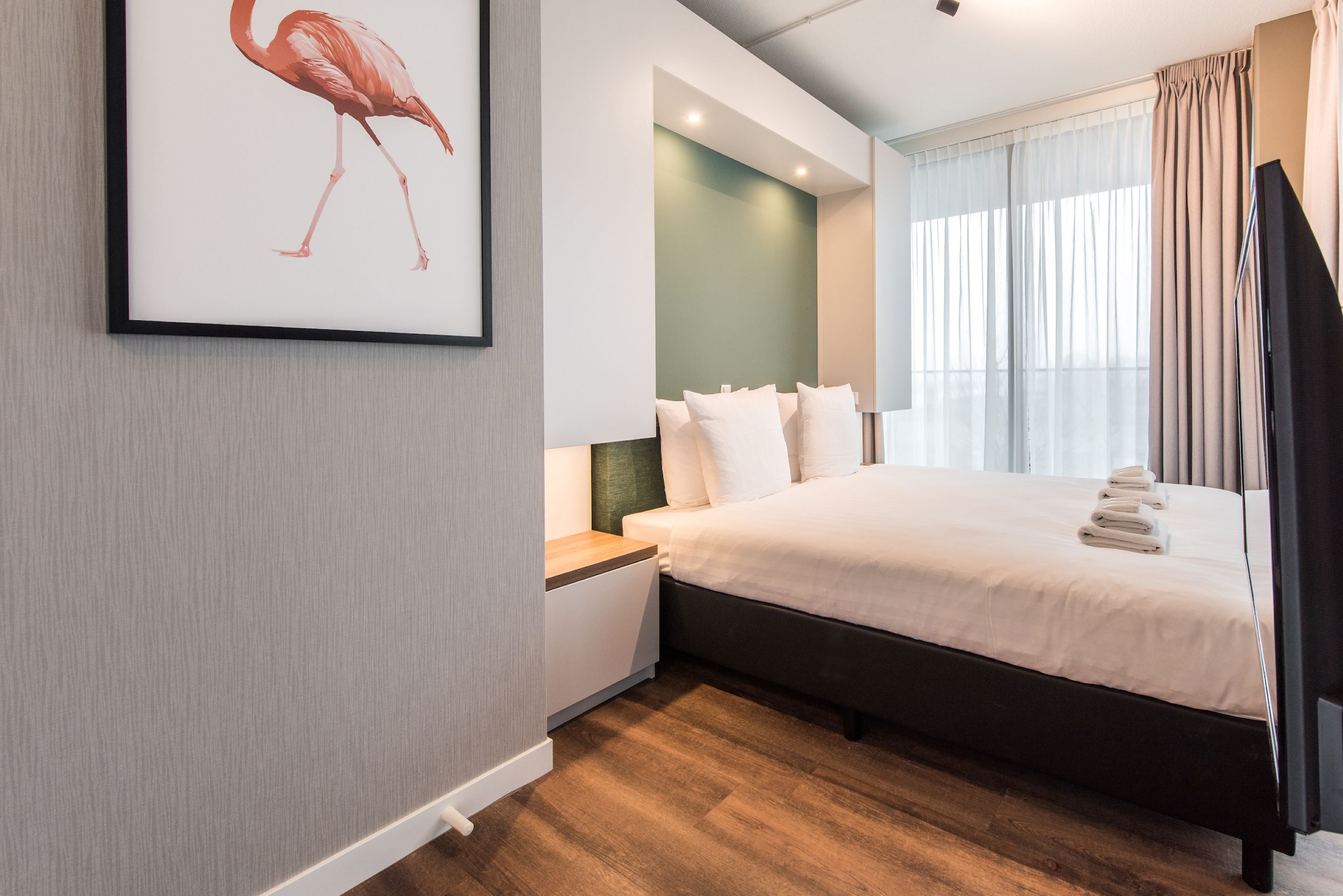 Una habitación de hotel moderna con una cama grande, una mesita de noche, una obra de arte enmarcada de un flamenco y una ventana con cortinas.
