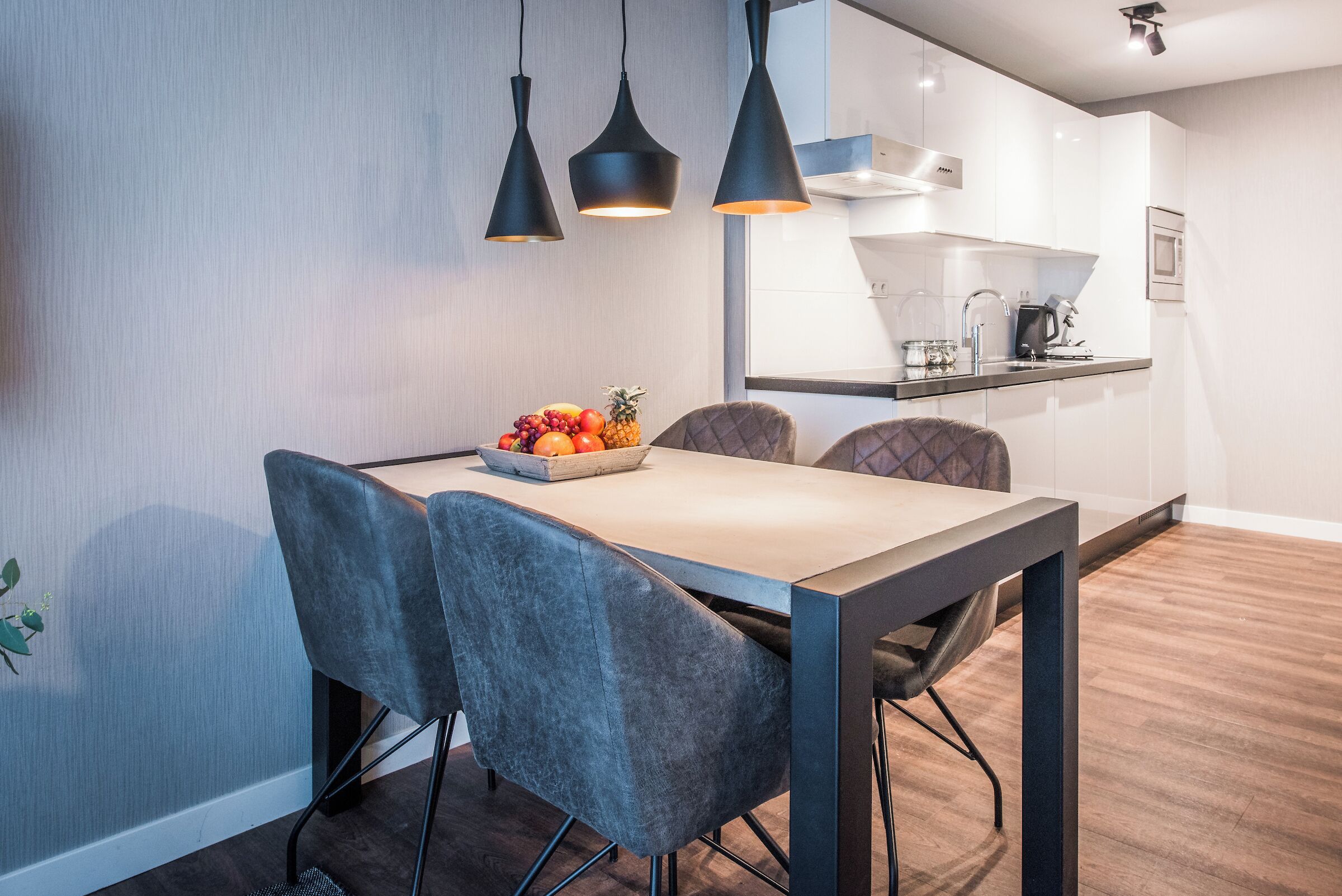 Et moderne spiseområde med et træbord, grå polstrede stole og en skål med frugt.