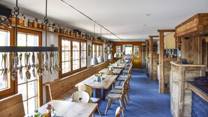 Een lange eetruimte met houten tafels en stoelen, een hangende rek voor keukengerei en een keuken met houten kasten.