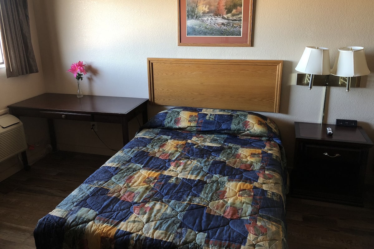 A hotel room with a bed, a desk, a chair, a lamp, and a painting on the wall.
