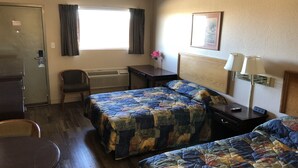 A hotel room with two beds, a desk, a chair, a small table, a window with curtains, a radiator, and a lamp.