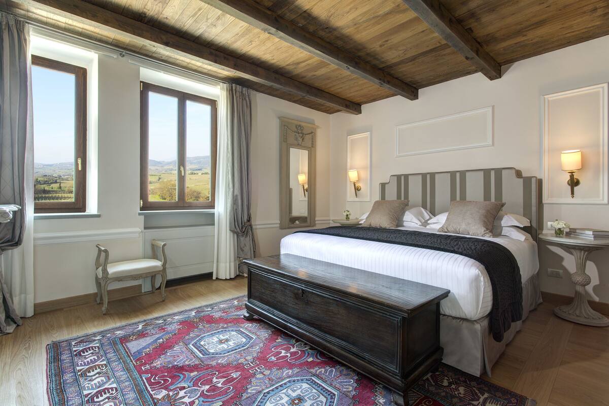 A bedroom with a large bed, a wooden bench, a patterned rug, and a view of the countryside through the windows.
