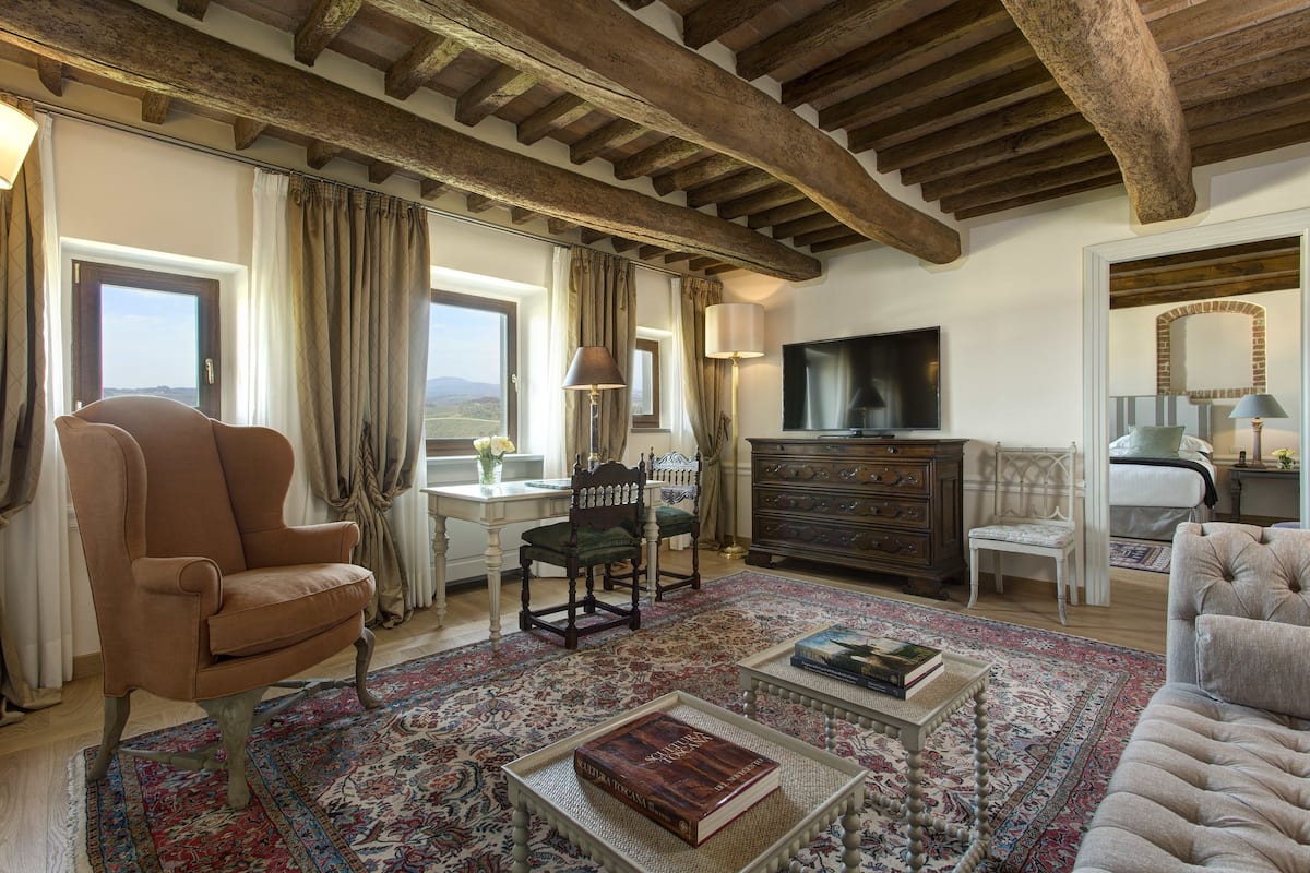 A spacious living room with a high ceiling, wooden beams, a large window with a view, a television, and a fireplace.