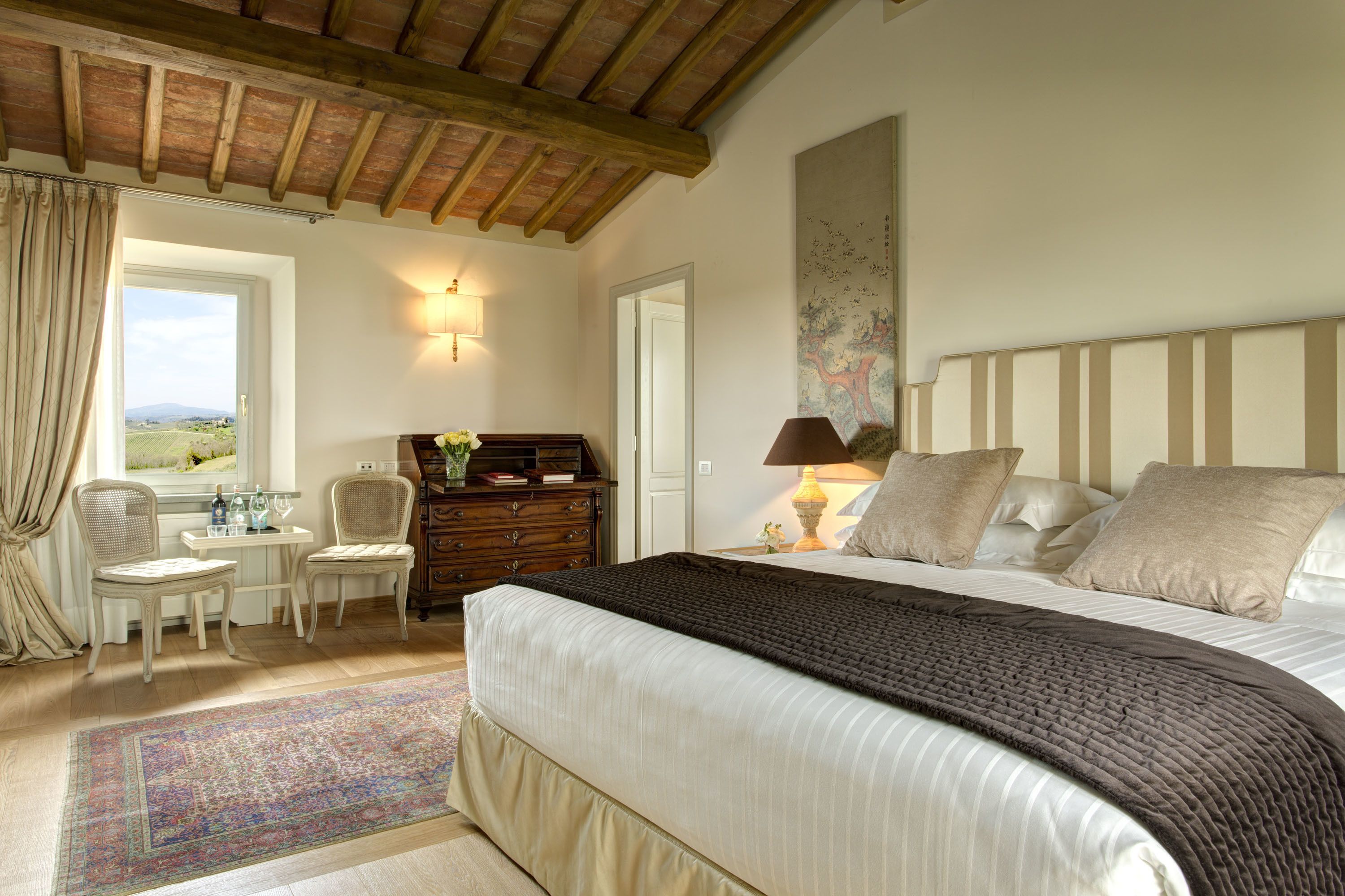 A bedroom with a large bed, a wooden ceiling, a desk with chairs, a dresser, and a window with curtains.