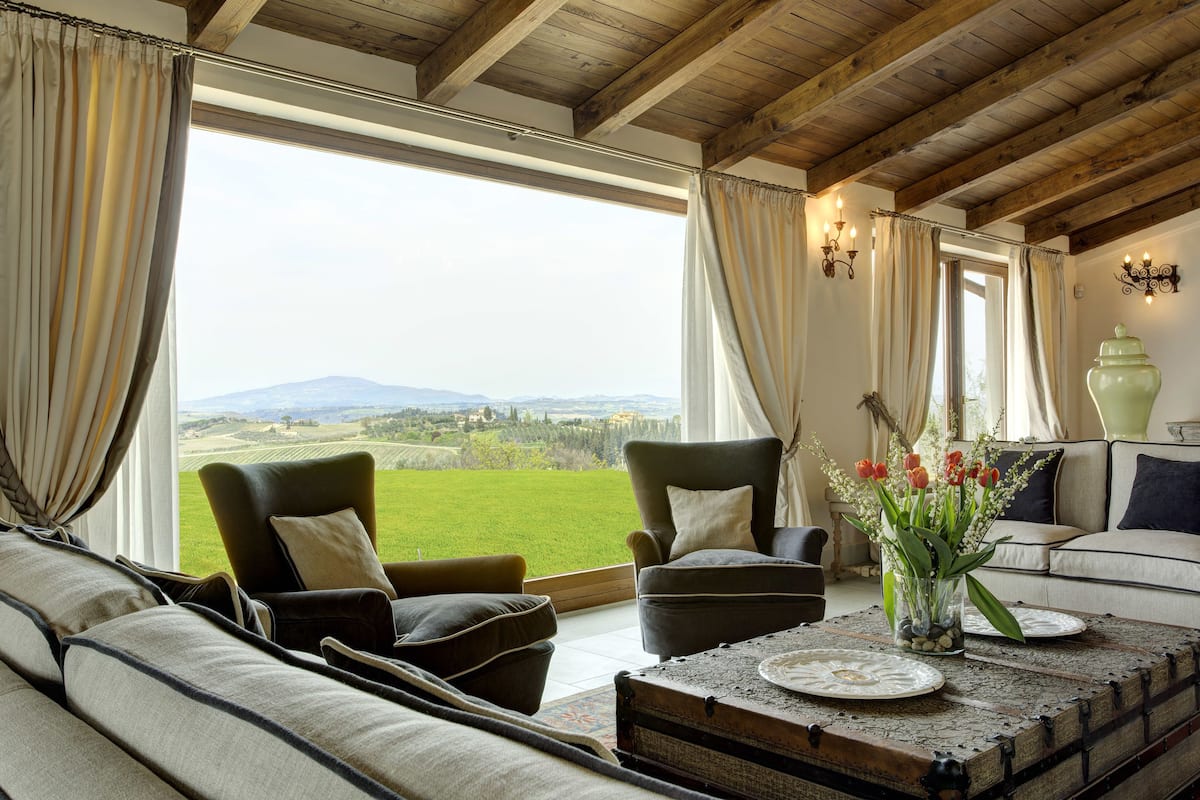 A spacious living room with a large window offering a view of the countryside. There are two armchairs, a sofa, and a coffee table with a vase of flowers.