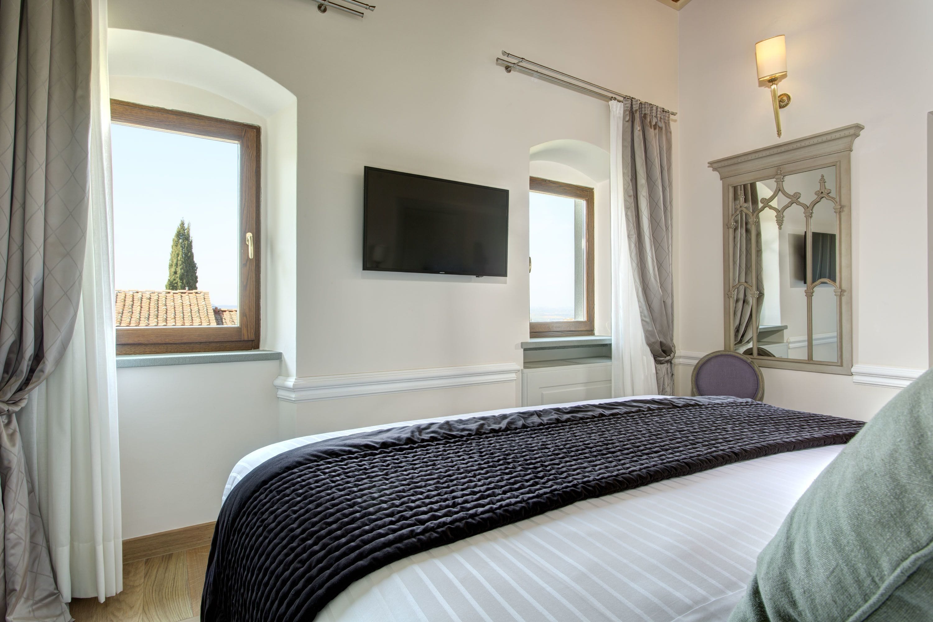 Habitación de hotel con una cama grande, un televisor de pantalla plana montado en la pared, una ventana con cortinas y un espejo con marco decorativo.