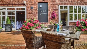 An outdoor dining area with wicker chairs and a table set for two, a blackboard with "Herzlich willkommen" written on it, and a brick building with large windows. - Hotel Hollenstedter Hof (Hollenstedt)