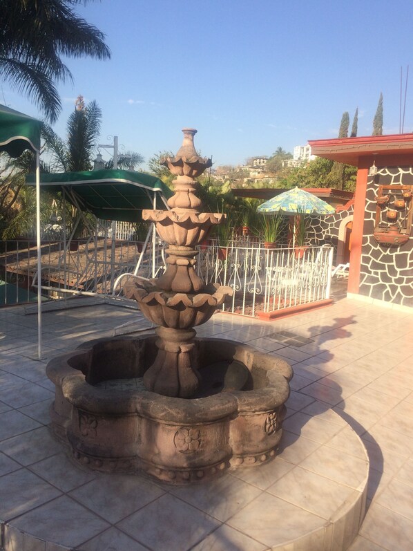Una fuente de piedra con diseño escalonado, rodeada por un patio con baldosas y una cerca de metal.