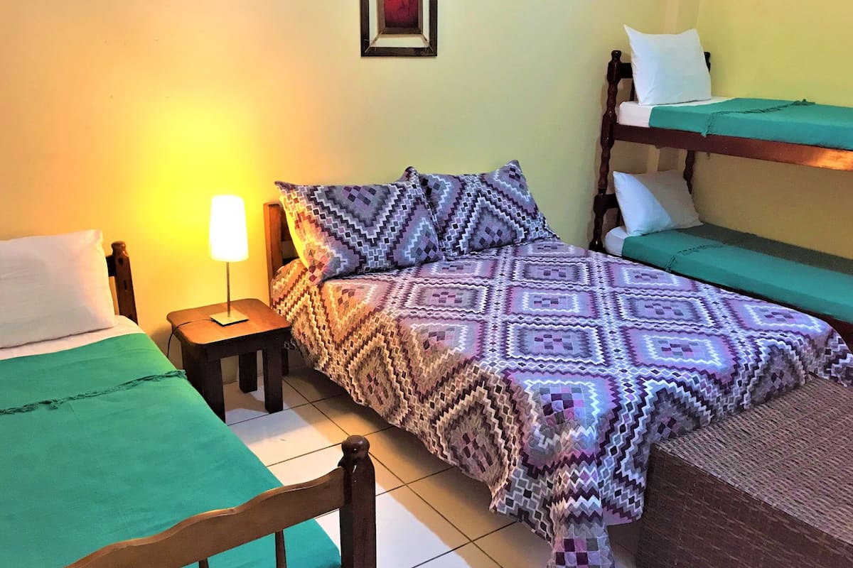 Une chambre avec deux lits : l’un est recouvert d’une couverture et de coussins à motifs, et l’autre d’un couvre-lit vert uni. Une petite table avec une lampe se trouve à côté du premier lit.
