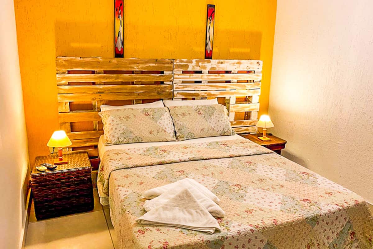 Un lit avec une couvre-lit à motifs floraux et des oreillers, une tête de lit en bois, deux lampes de chevet et une petite table de chevet avec une lampe et un panier.