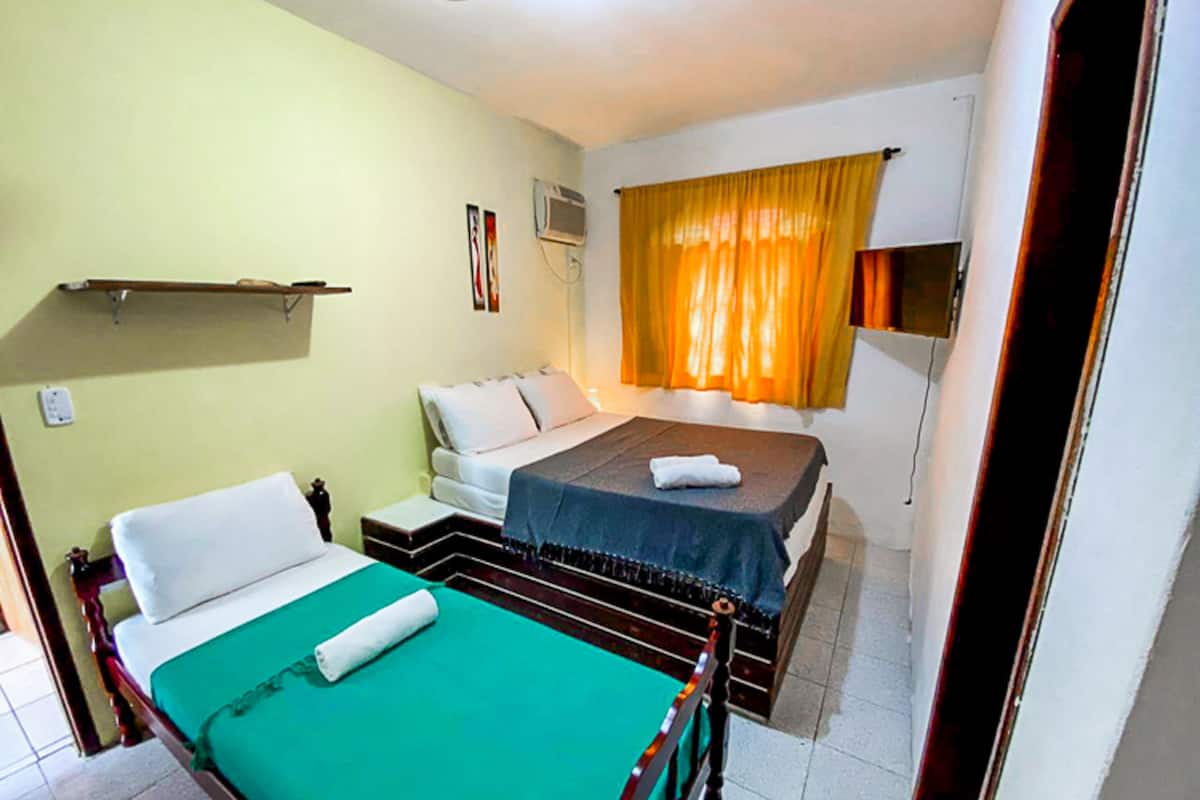 Une petite chambre propre, avec deux lits, une table de chevet, une étagère et une fenêtre avec des rideaux.