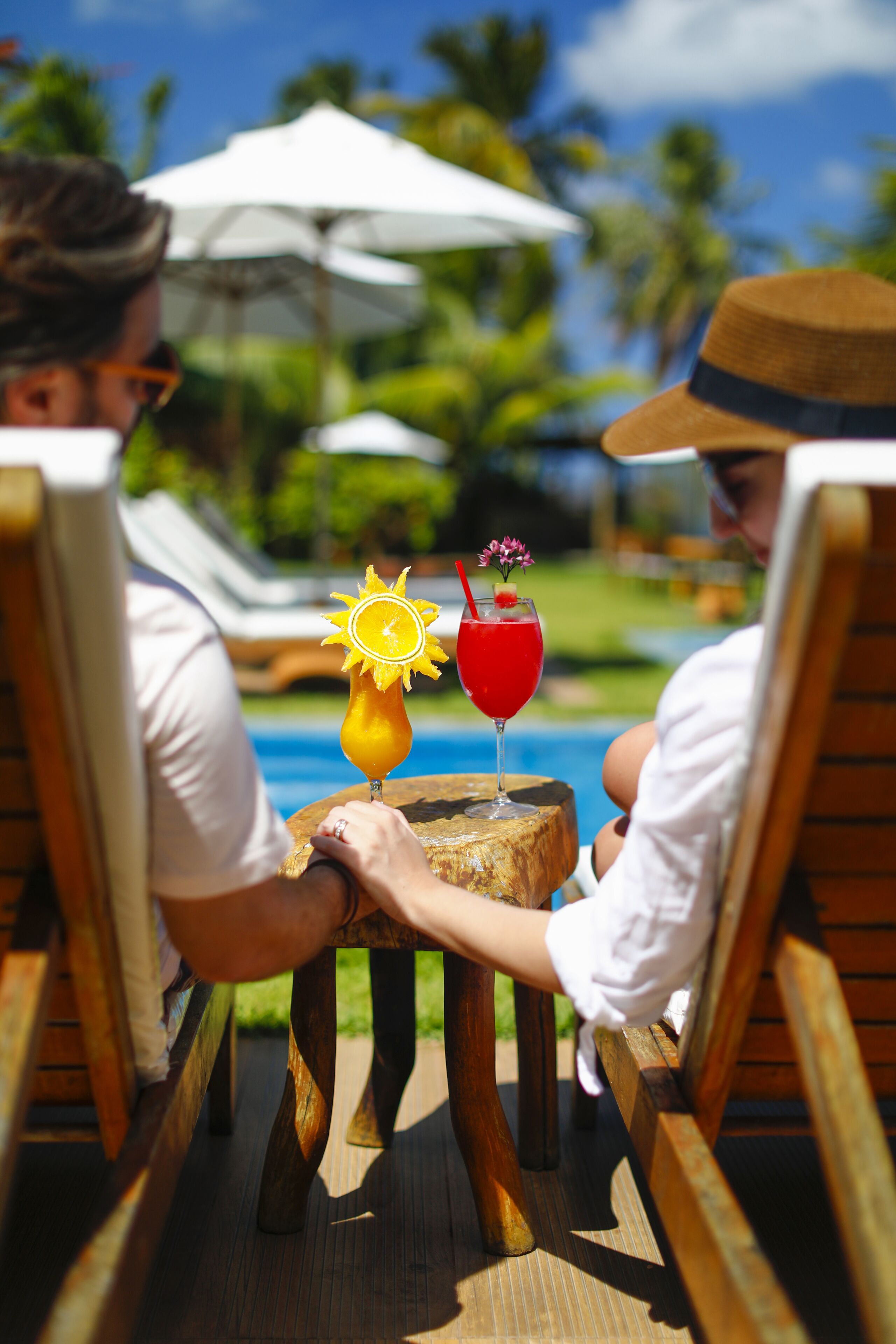 Two people sitting on wooden lounge chairs by a poolside, with a tropical setting in the background.