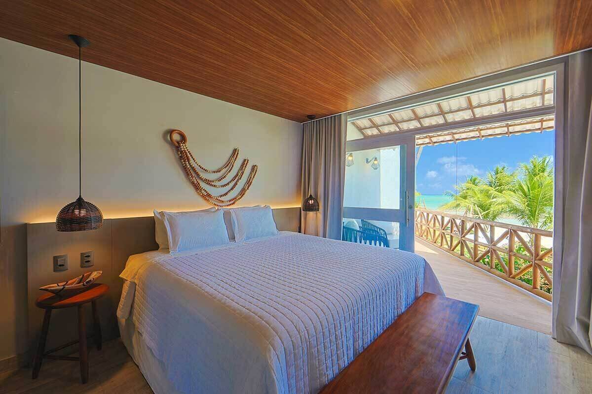 Ein Schlafzimmer mit einem großen Bett, einer hölzernen Bank, einer Pendelleuchte und Blick auf das Meer durch eine Schiebetür.