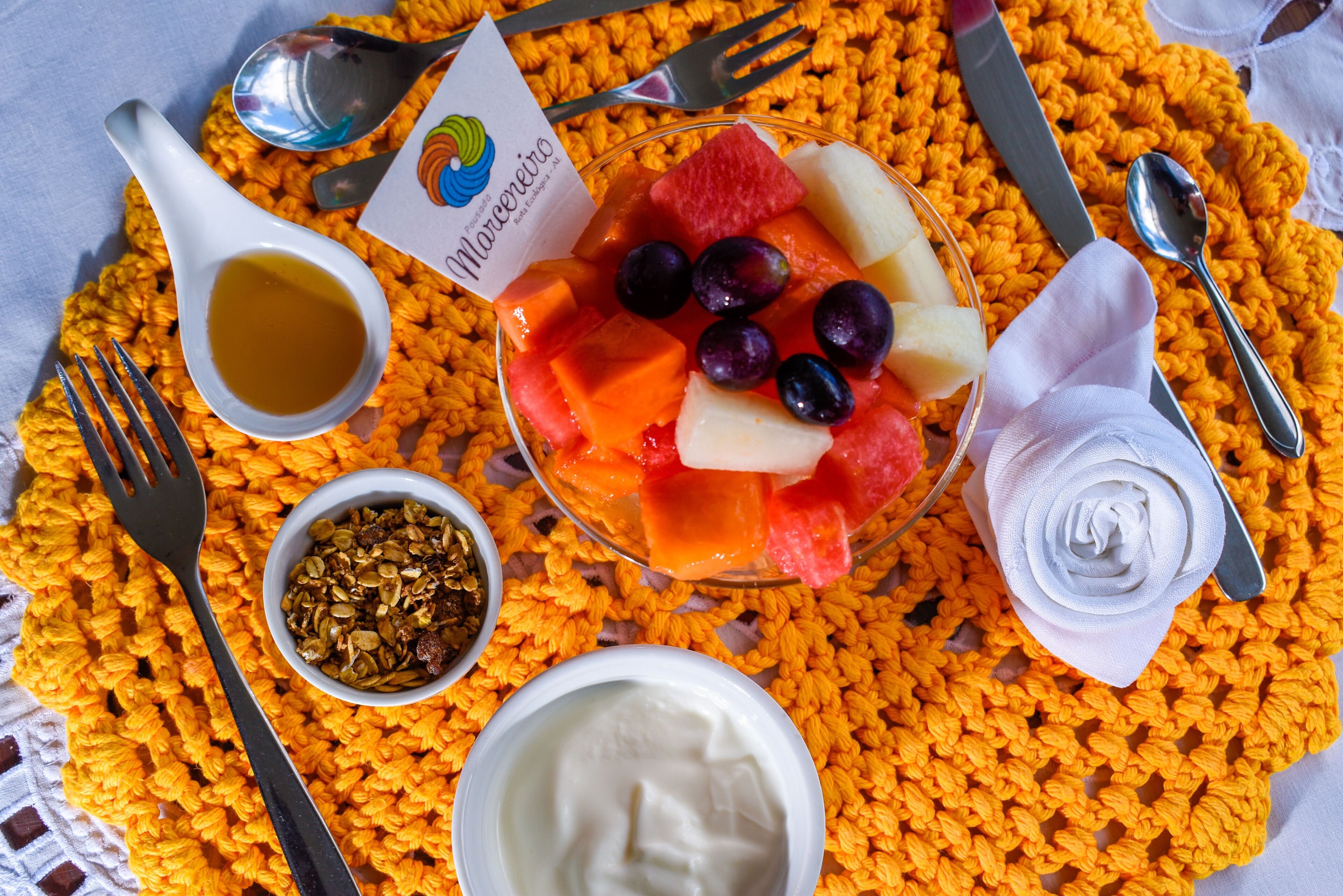 A breakfast setting with a fruit bowl, yogurt, granola, and a napkin on a yellow knitted placemat.