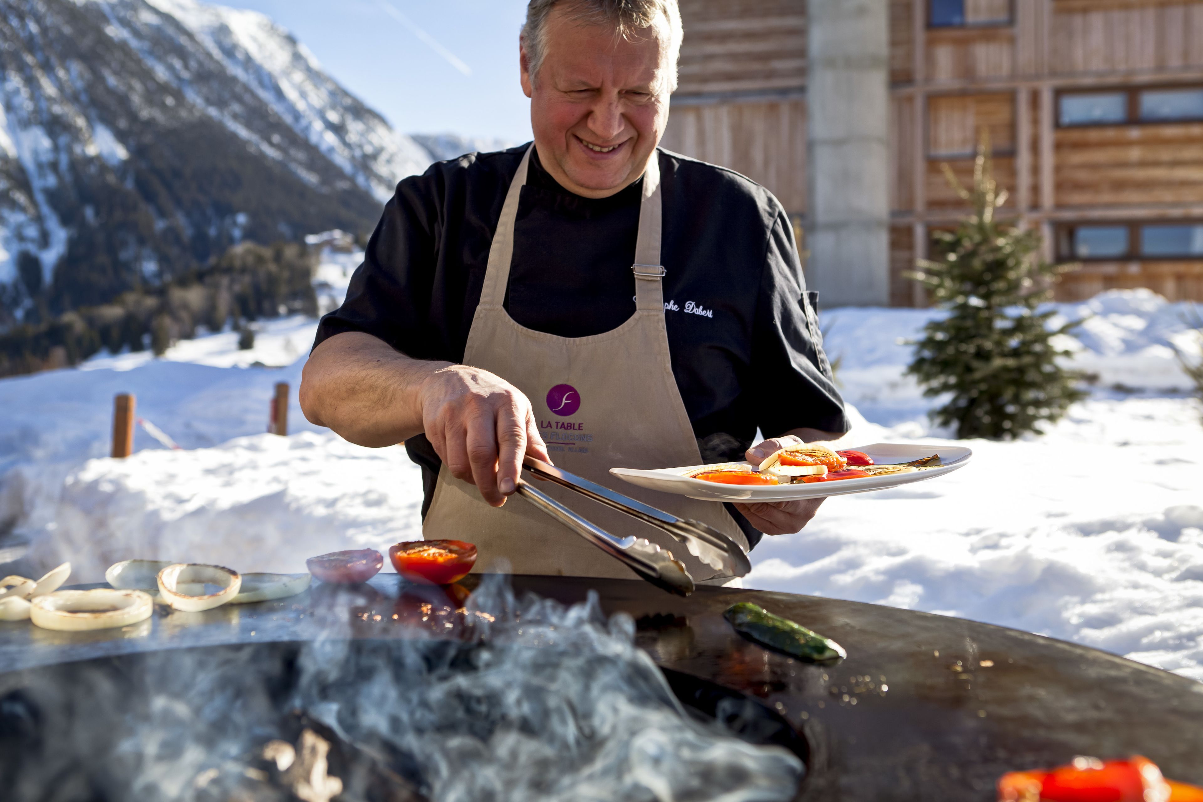 Une personne cuisine à l’extérieur sur un barbecue, avec de la neige en arrière-plan et des montagnes.