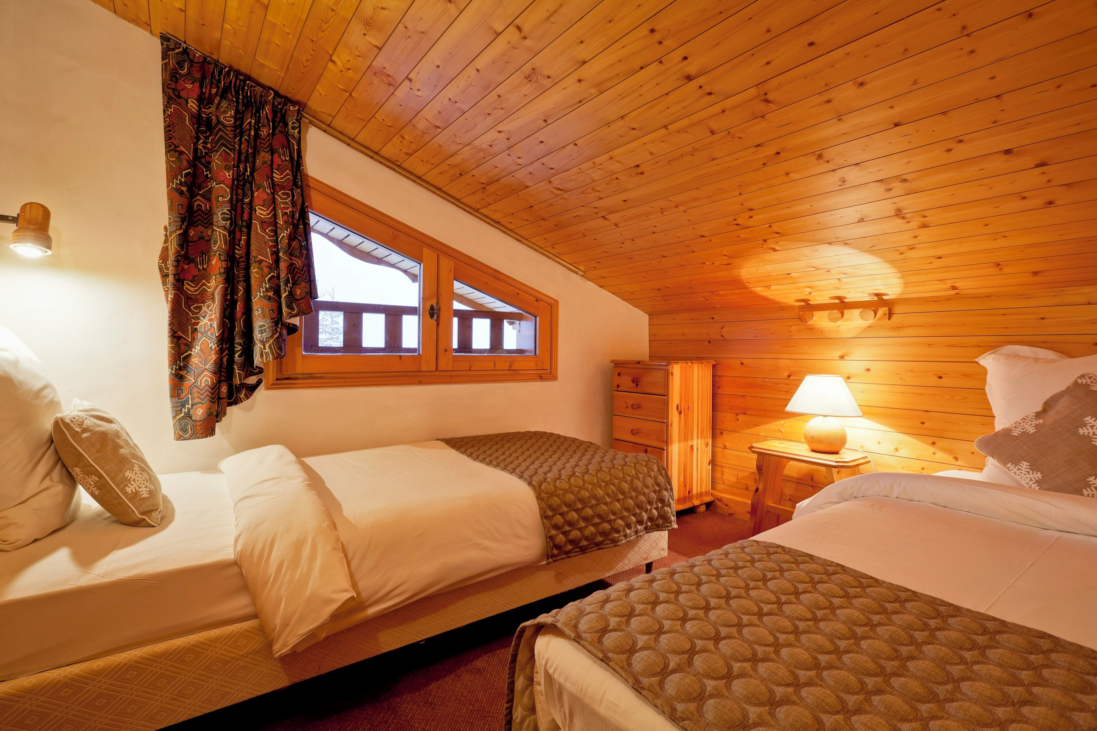 Een knus ogende kamer met houten plafonds en wanden, twee bedden met beddengoed met een patroon, een raam met gordijnen en een bedlampje.