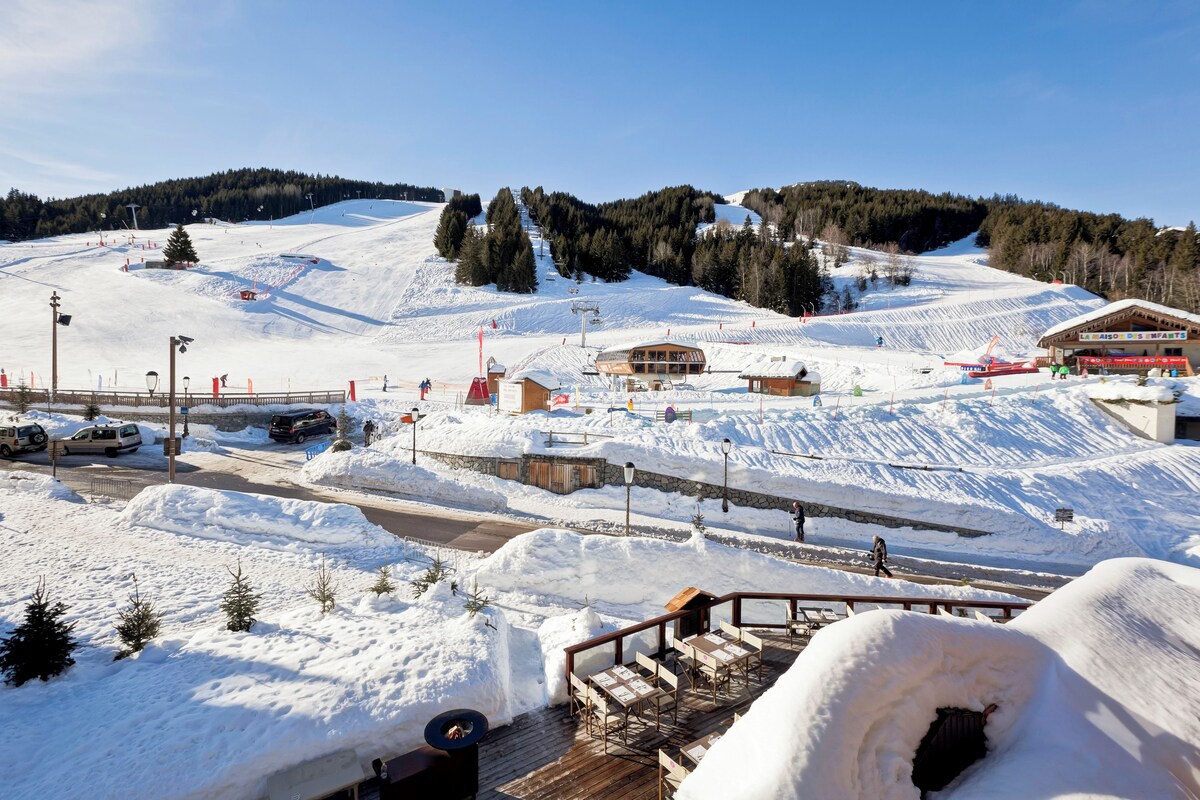 A ski resort with a wooden deck, snow-covered landscape, and a building with a ski lift in the background.