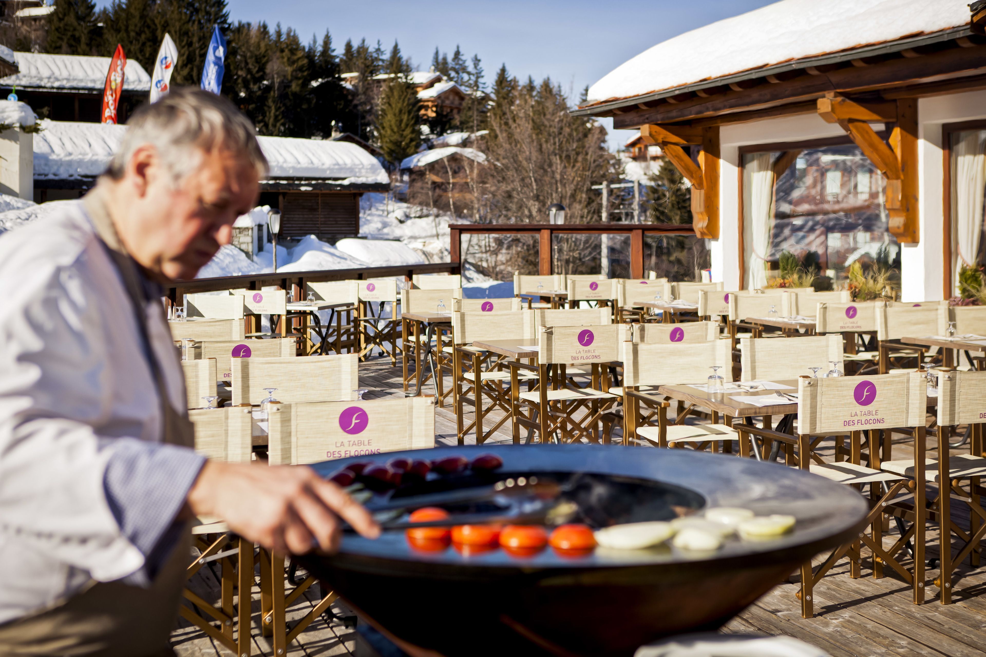 An outdoor dining area with wooden tables and chairs, a grill with vegetables, and a mountainous backdrop.