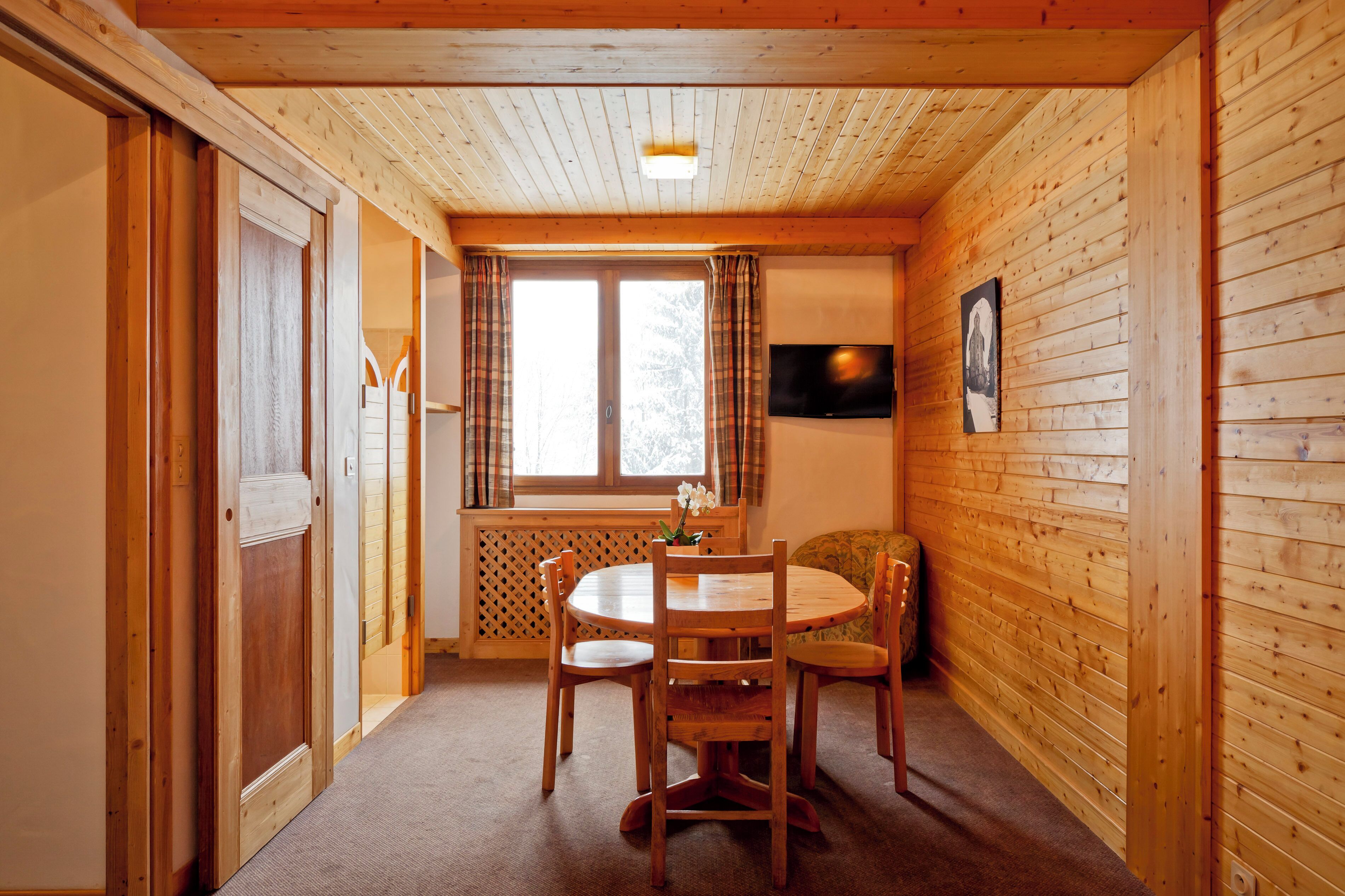A wooden-paneled room with a round wooden table and four chairs, a window with curtains, and a television on the wall.